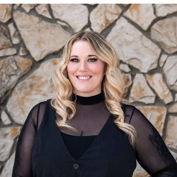 Smiling woman with blonde curls in black sheer-sleeved top by a stone wall
