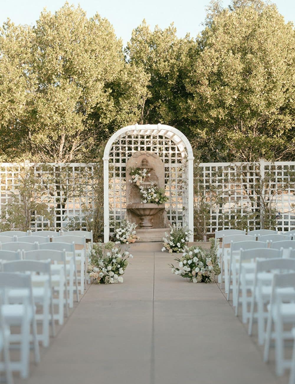 Outdoor ceremony aisle with white chairs facing a lattice arch and stone fountain