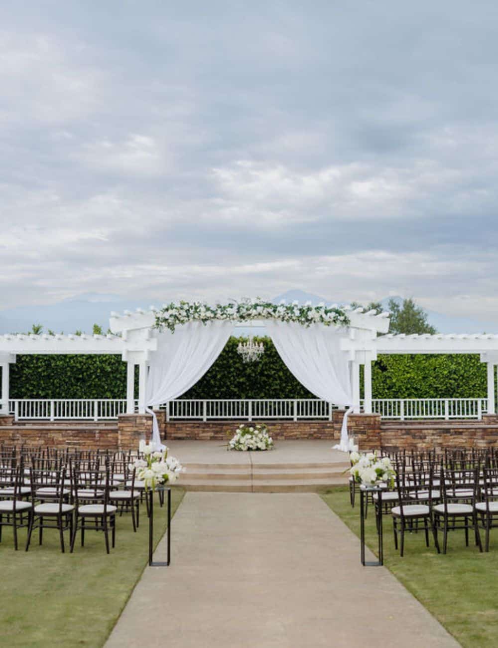 White pergola with draping and florals framing a ceremony aisle lined with chairs