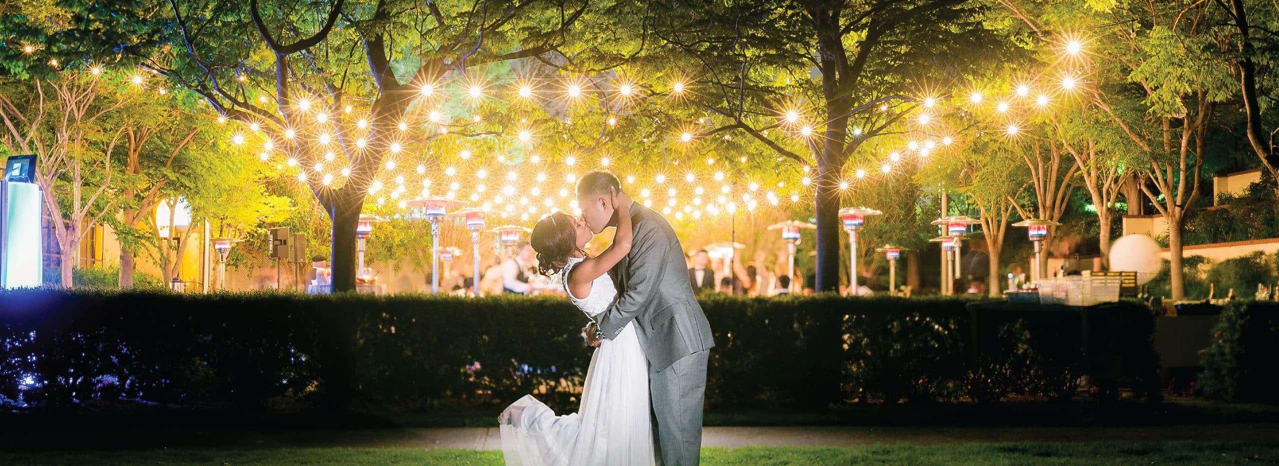 Couple kissing on a lawn beneath strings of glowing cafe lights at Mountaingate