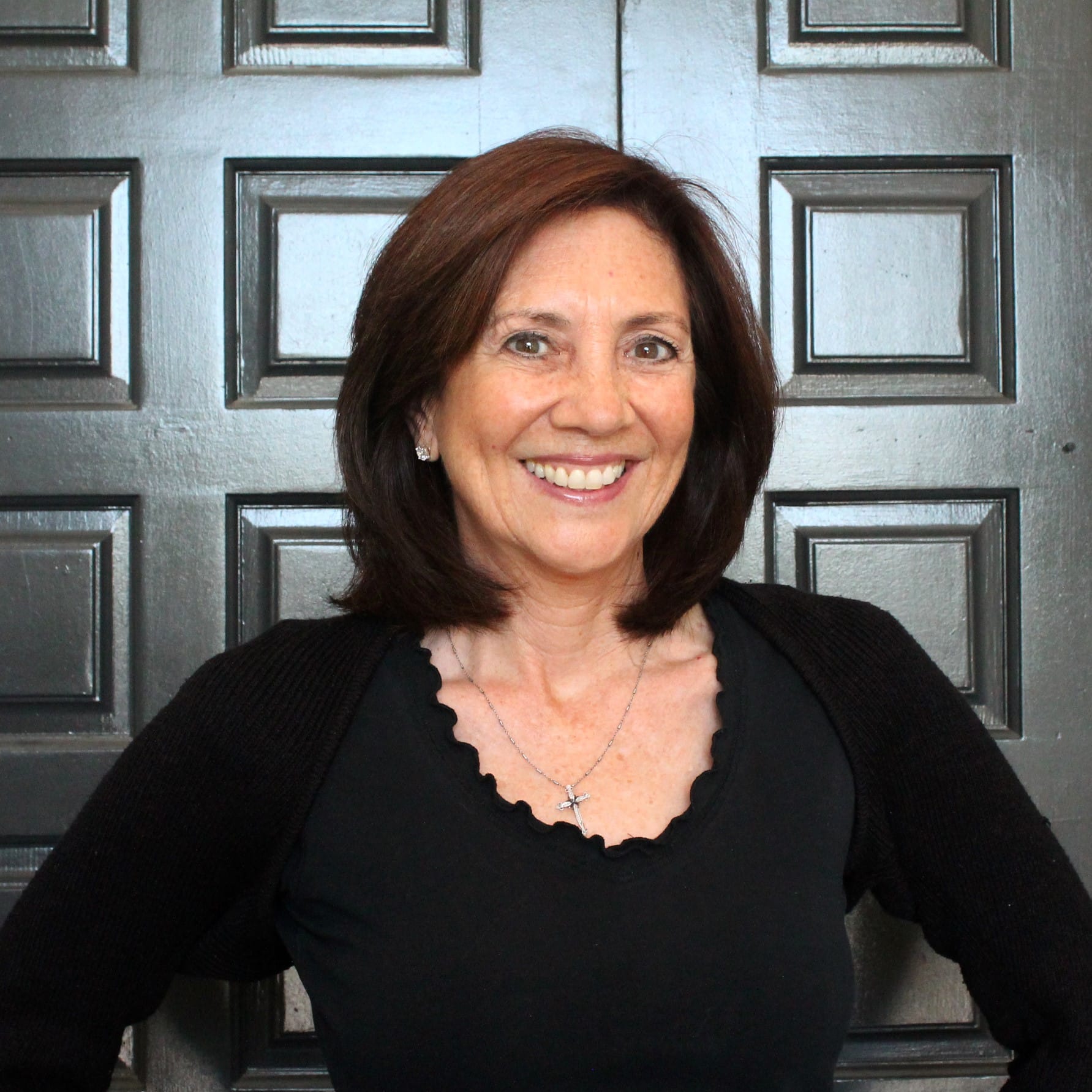 Rochelle, smiling in a black top in front of a dark paneled door
