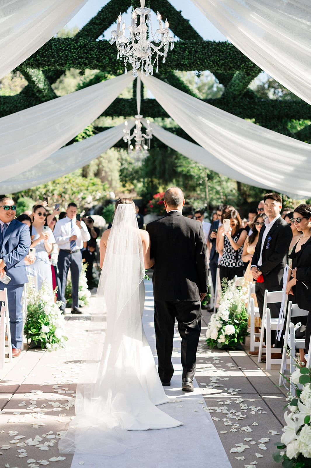 Bride escorted down a draped outdoor aisle under crystal chandeliers