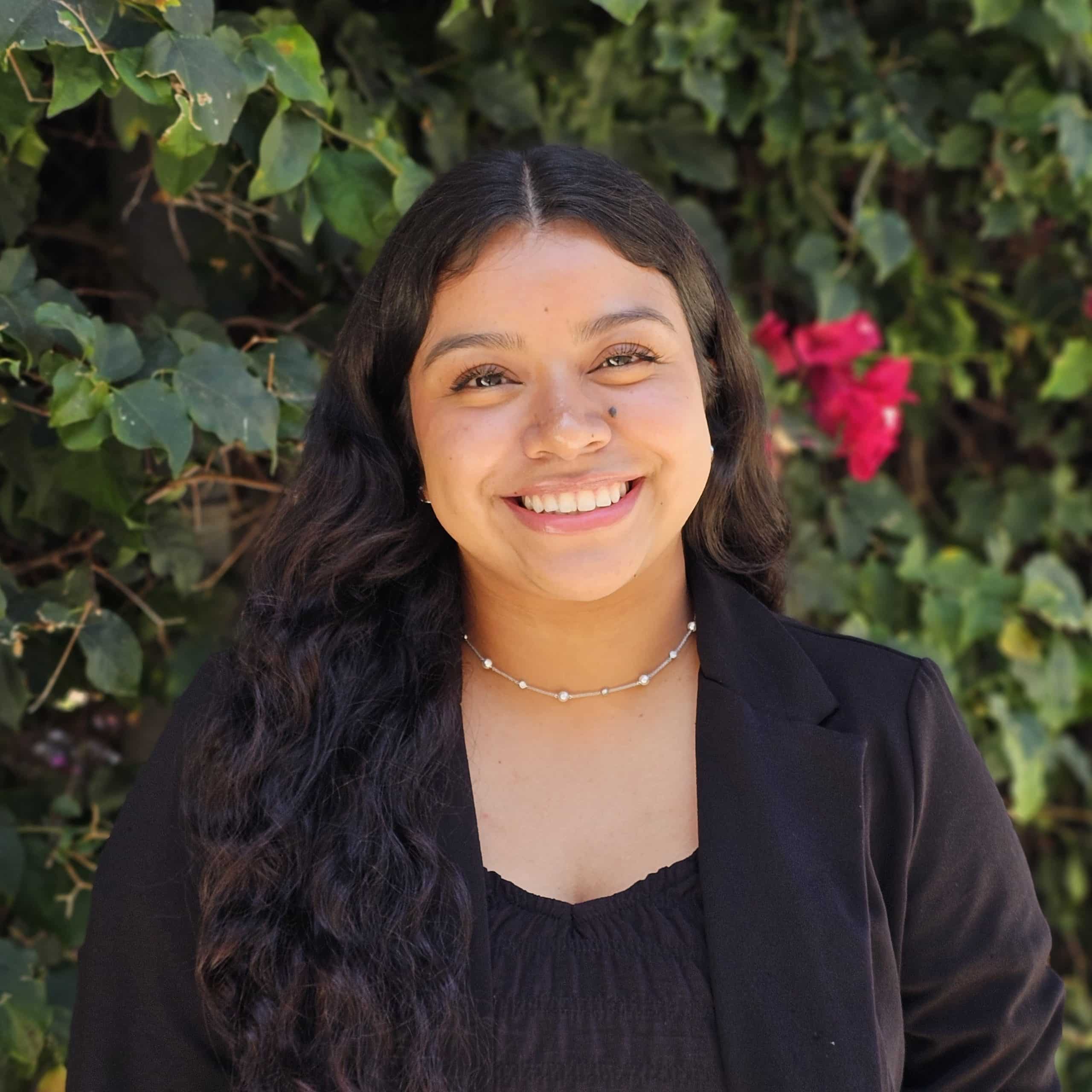 Grecia Carmona, smiling in a black blazer in front of flowering greenery